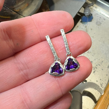 trillion-shaped purple amethyst gemstone earrings held between fingers with a workshop background