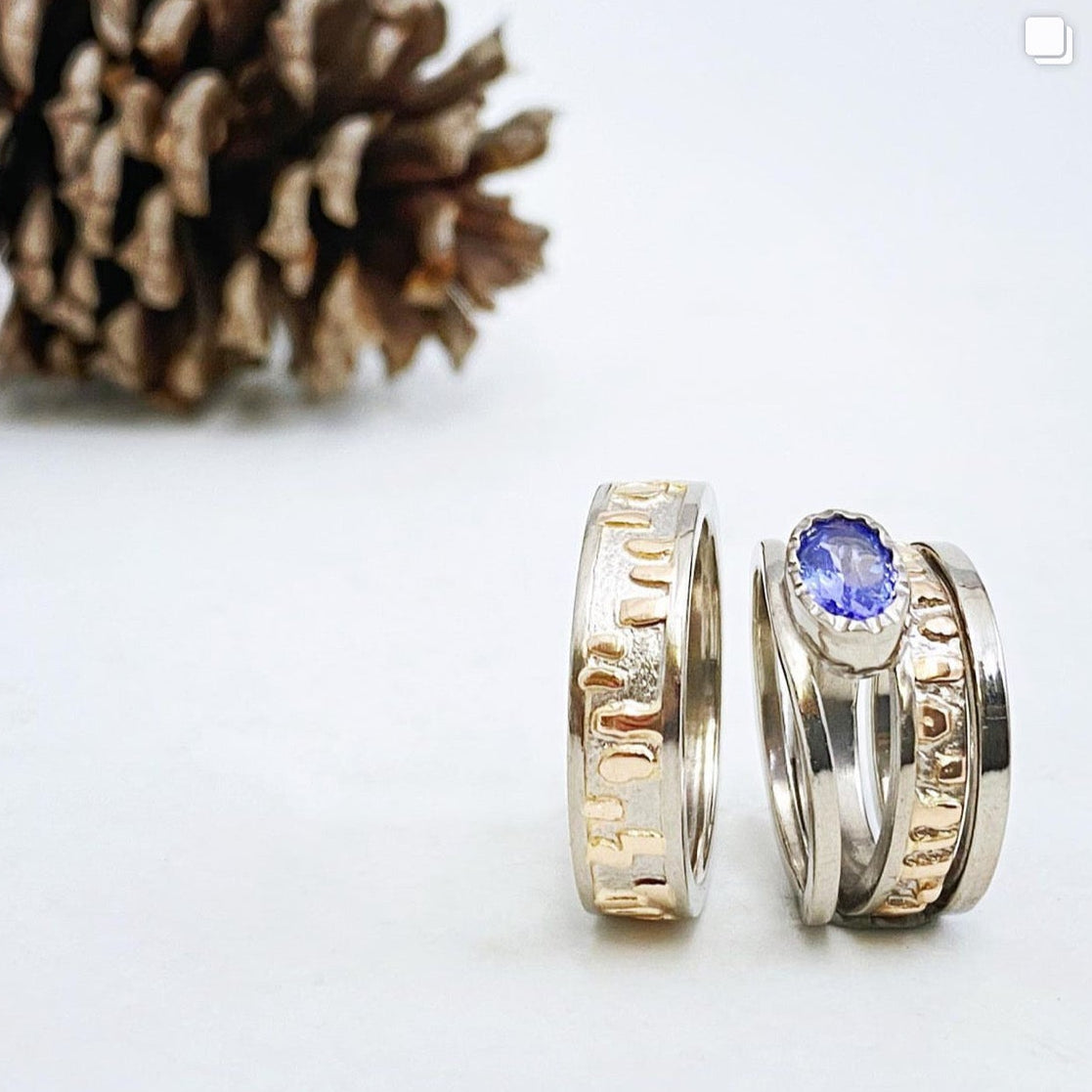 Two white and yellow gold rings with engraved designs and a blue tanzanite gemstone next to a pine cone on a white background